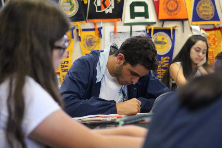 Senior Samuel Metta writes down a list of due dates told by IB Literature HL 2 teacher Eric Helle during his fifth period class. The background shows an array of college benners that have been in Helles classroom since her began teaching.